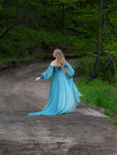 One Blonde Haired Woman Wearing Long Flowing Blue Gown In Green Meadow. Magical, Fairytale Concept.	