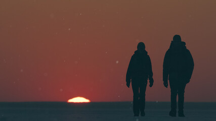 The two travelers standing through the snow field against beautiful sunset