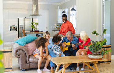 Wall Mural - Multiracial senior man playing guitar while sitting with happy multigeneration family on sofa