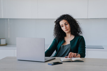 Exhausted curly hispanic young woman in green clothes sitting at desk with laptop and diary. Sad caucasian businesswoman received  bad news during video call. Beautiful arabic entrepreneur tired.
