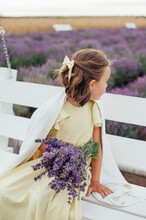 Pretty Little Girl Is Looking Back Wearing Yellow Dress And A Bow In Hair With A Bouquet Of Purple Flowers In Hands Is Sitting On A Swing Among Lavender Field
