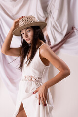 Wall Mural - Portrait of beautiful woman in straw hat and white dress posing isolated over textile background. Romantic outfit.