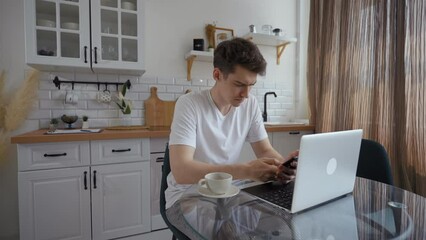 Young man types message on phone sitting near laptop at glass table in modern kitchen. Busy guy distracts on gadget while working online at home
