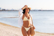 © Syda Productions - people, summer holidays and leisure concept - happy young woman in bikini swimsuit, white shirt and straw hat with bag walking along beach