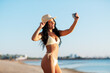 © Syda Productions - people, summer and swimwear concept - happy smiling young woman in bikini swimsuit and straw hat taking selfie with smartphone on beach