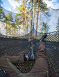 A man is lying on net hammock in forest. First-person view of the legs.