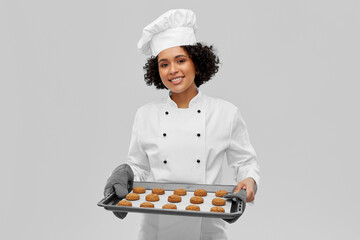 cooking, culinary and bakery concept - happy smiling female chef or baker in white toque and jacket holding baking tray with oatmeal cookies over grey background