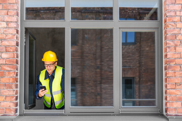 Poster - construction business and building concept - smiling male builder in helmet and safety west with smartphone at open window