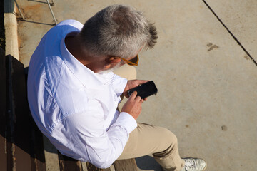 Handsome mature man with beard and grey hair is sitting thoughtfully on a park bench while consulting his mobile phone. The man is dressed in beige trousers, white shirt. Concept of old age.