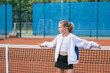 Portrait of a girl in sportswear standing near tennis court net. Junior tennis player on an outdoor tennis court.