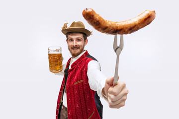 Wall Mural - Joyful man wearing traditional fest Bavarian or German outfit with big beer glass showing fried sausage on fork. Oktoberfest, party, holiday, ad, sales concept