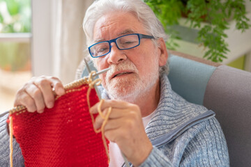 Canvas Print - Portrait of senior man checking knitted wool with needle sitting on sofa at home. Happy caucasian man knit wool while relaxing at home.