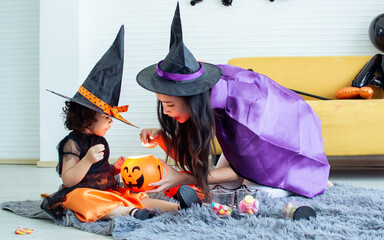 Asian beautiful woman or mother and little Caucasian sweet girl wearing witch costume with hats, playing pumpkin, trick or treat with fun, smiling with happiness, celebrating Halloween party at home.