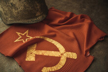 A Red Flag With A Yellow Hammer And Sickle. A Soldier's Helmet On The Red Flag Of The Soviet Union. The Flag Of The Communist Party On A Wooden Table.