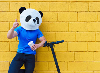 Man with panda mask making ok gesture leaning on electric scooter with drink in other hand.