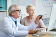 © Mediaphotos - Serious gray-haired doctor in lab coat sitting at table and pointing at computer monitor while showing online medical records to patient