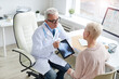 © Mediaphotos - Serious aged doctor in white coat sitting at table and using tablet while showing online x-ray image of spine to patient
