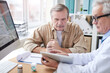 © Mediaphotos - Serious male doctor sitting at desk with computer and showing medical record on tablet to aged patient
