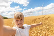 Portrait of little cute beautiful blond caucasian girl enjoy having fun taking selfie photo phone camera walking at wheat field against wind mill turbine farm. Children blogging and video streaming