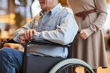 Close-up Of Female Volunteer In Dress Pushing Aged Man In Wheelchair While Working With Handicapped Man In Nursing Home