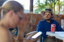 Young Bloke With Cowboy Hat, Beer, Sunnies With Blurry Foreground Of Girl On Phone