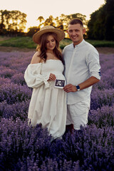 Wall Mural - portrait of a young beautiful pregnant couple on a lavender field at sunset holding first ultrasonography image. Happy family concept