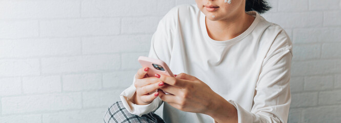 Wall Mural - girl is texting on a smartphone while sitting on a bed at home. copy space. teenage girl with problematic skin, with a therapeutic cream applied.