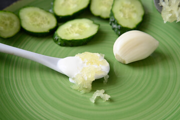 squeezed garlic and fresh cucumbers cut into slices on a green plate. white spoon of sour cream.