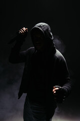 Wall Mural - Silhouette of african american man in hood and gloves holding knife on black background with smoke