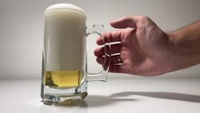 There Is A Glass Of Cold, Pale Beer On A White Background. There Is Beer In The Glass And Foam From It. A Man's Hand Slowly Reaches For The Glass And Takes It Away.