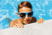 Cute Boy In Sunglasses Leaning On The Edge Of The Pool During Summer Vacation. The Boy Looks At The Camera In The Pool