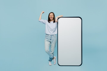 Wall Mural - Full body young woman she 20s wear casual blouse big huge blank screen mobile cell phone with workspace copy space mockup area do winner gesture isolated on pastel plain light blue background studio.