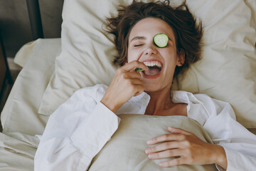 Wall Mural - Young fun woman wear white shirt pajama she lying in bed with mask on eyes eat biting cucumber slices rest relax spend time in bedroom lounge home in hotel room wake up Skin care wellbeing concept.