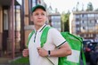 © Евгений Шемякин - portrait of young handsome happy courier guy, food delivery man with green thermo box for food delivering food outdoors in the yard on buildings background at summer day in cap and uniform.