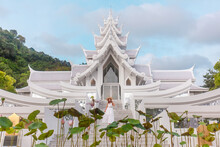 Traveler Woman In Dress Walking In Front Of White And Silver Temple In Thai Style. Travel And Vacation In Asia. Buddhist Temple On Phuket Island In Thailand With Blooming Lotus In Foreground.