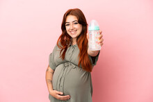 Young Redhead Caucasian Woman Isolated On Pink Background Pregnant And Holding A Feeding Bottle