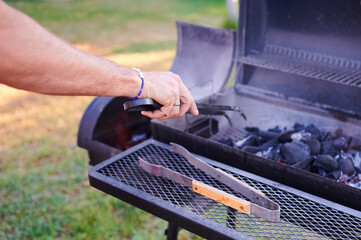 Unrecognizable man prepares the burning coal with which he is going to cook a barbecue on a grill in the backyard of the house using barbecue utensils.