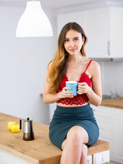 Wall Mural - Beautiful woman sitting on countertop at kitchen and holding cup of coffee