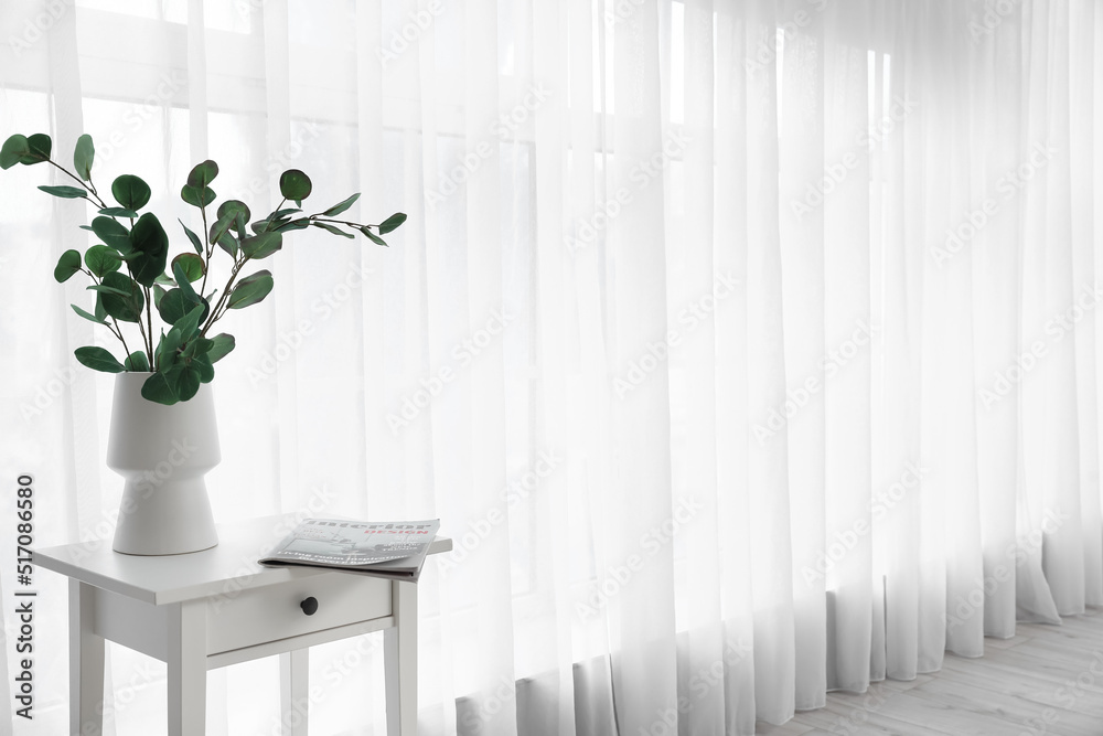 Vase with eucalyptus branches on table near light curtain