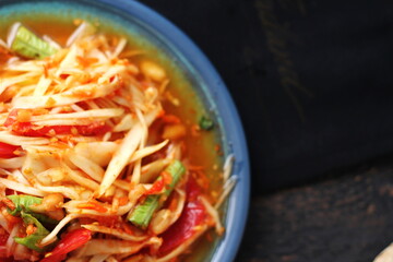 Spicy papaya salad served in a blue dish with sticky rice in a bucket and long beans, dried chilli and vegetables.