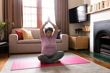 Biracial Mature Woman With Arms Raised And Eyes Closed Meditating While Sitting On Mat At Home