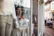 © Caia Image - Female shop owner with tea looking out shop window