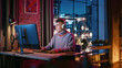 Young Handsome Man in Headphones Using Computer in Stylish Loft Apartment at Night. Creative Male Working from Home, Browsing Internet and Social Media. Urban City View from Big Window.