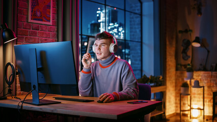 Leinwandbilder - Young Handsome Man in Headphones Using Computer in Stylish Loft Apartment at Night. Creative Male Working from Home, Browsing Internet and Social Media. Urban City View from Big Window.