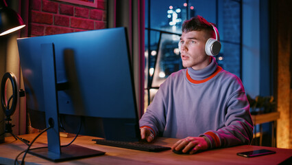 Wall Mural - Young Handsome Man in Headphones Using Computer in Stylish Loft Apartment in the Evening. Creative Male Smiling, Working from Home, Browsing Videos on Social Media. Urban City View from Big Window.