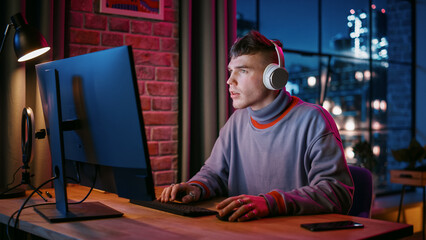 Wall Mural - Young Handsome Man in Headphones Using Computer in Stylish Loft Apartment in the Evening. Creative Male Smiling, Working from Home, Browsing Videos on Social Media. Urban City View from Big Window.