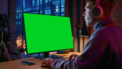 Fototapete - Young Handsome Man in Headphones Working from Home on Desktop Computer with Green Screen Mock Up Display. Creative Male Checking Social Media, Browsing Internet. Urban City View from Big Window.