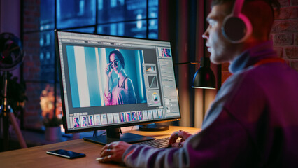 Fototapete - Young Creative Man in Headphones Using Computer in Stylish Loft Apartment in the Evening. Graphic Designer Working from Home, Editing Fashion Photo for Online Store. Urban City View from Big Window.