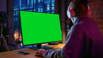 Young Handsome Man in Headphones Working from Home on Desktop Computer with Green Screen Mock Up Display. Creative Male Checking Social Media, Browsing Internet. Urban City View from Big Window.