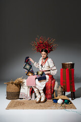 Wall Mural - full length of brunette ukrainian woman in traditional clothes and red floral wreath holding knitted scarf on dark grey.